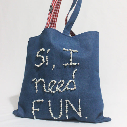 Blue tote bag with 'Si, I need fun' text and checkered handle on a white background