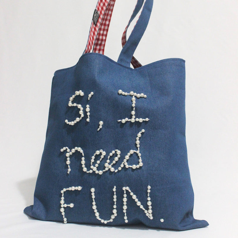 Blue tote bag with 'Si, I need fun' text and checkered handle on a white background